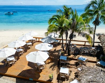 Beachside deck with white umbrellas and wooden tables, surrounded by palm trees. Overlooks turquoise ocean, conveying a serene, tropical atmosphere.