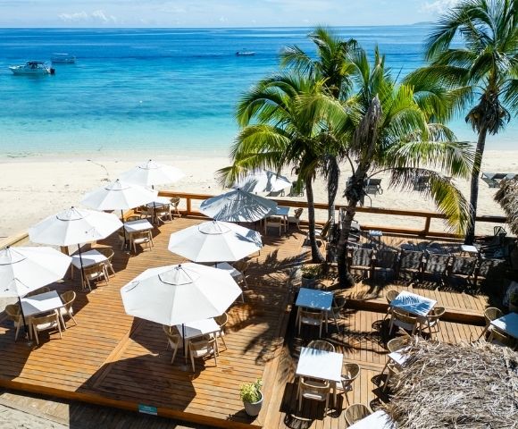 Beachside deck with white umbrellas and wooden tables, surrounded by palm trees. Overlooks turquoise ocean, conveying a serene, tropical atmosphere.