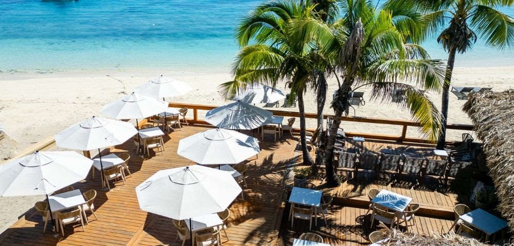 Beachside deck with white umbrellas and wooden tables, surrounded by palm trees. Overlooks turquoise ocean, conveying a serene, tropical atmosphere.