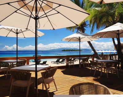 Beachside dining area with wooden tables, chairs, and white umbrellas. Overlooks a serene ocean and palm trees against a bright blue sky. Relaxing mood.