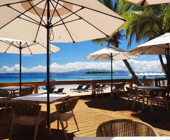 Beachside dining area with wooden tables, chairs, and white umbrellas. Overlooks a serene ocean and palm trees against a bright blue sky. Relaxing mood.