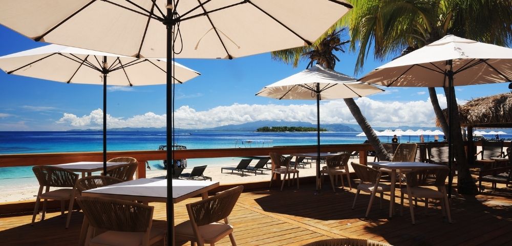 Beachside dining area with wooden tables, chairs, and white umbrellas. Overlooks a serene ocean and palm trees against a bright blue sky. Relaxing mood.