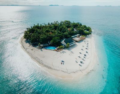 Aerial view of a small tropical island with lush greenery, surrounded by turquoise water. White sand beach features sun loungers and a swimming pool. Serene and idyllic.