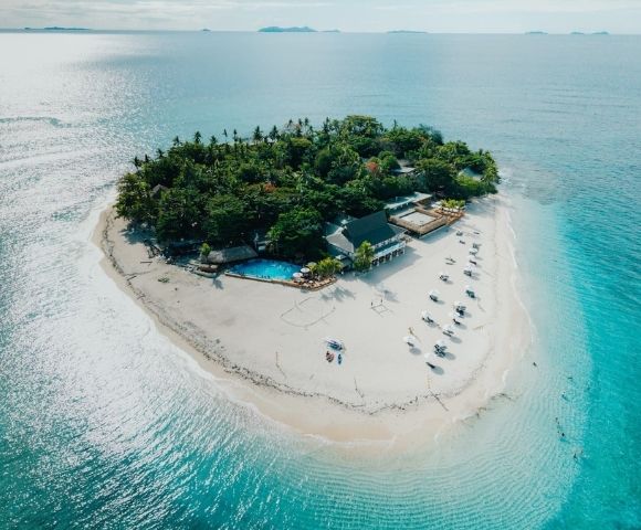 Aerial view of a small tropical island with lush greenery, surrounded by turquoise water. White sand beach features sun loungers and a swimming pool. Serene and idyllic.