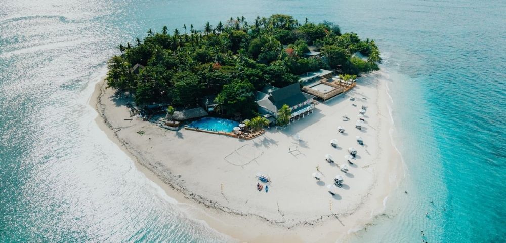 Aerial view of a small tropical island with lush greenery, surrounded by turquoise water. White sand beach features sun loungers and a swimming pool. Serene and idyllic.