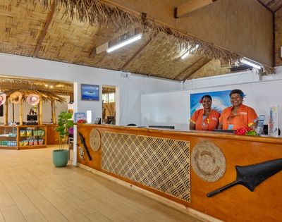 Reception area with two smiling staff in orange uniforms behind a wooden desk with decorative carvings. The room features straw ceiling accents.