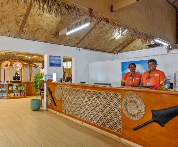 Reception area with two smiling staff in orange uniforms behind a wooden desk with decorative carvings. The room features straw ceiling accents.