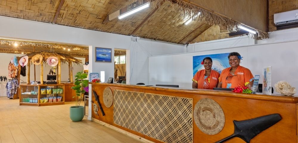 Reception area with two smiling staff in orange uniforms behind a wooden desk with decorative carvings. The room features straw ceiling accents.