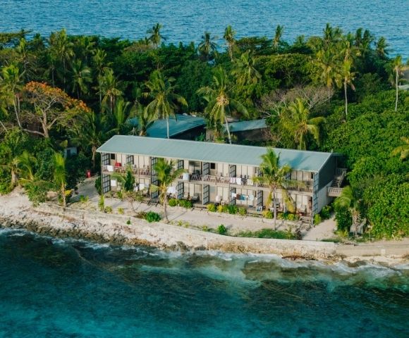 Coastal resort with a long building surrounded by lush palm trees sits on a rocky shore. The blue ocean in the foreground conveys a tropical, tranquil vibe.