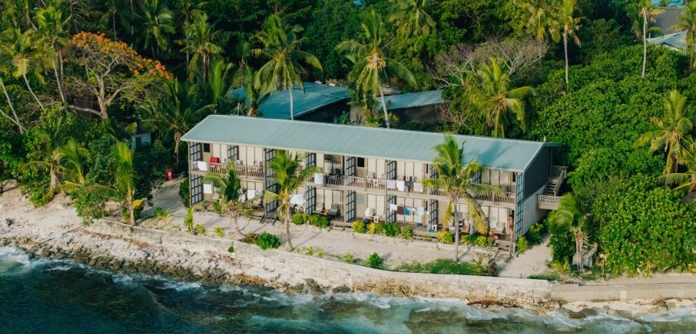 Coastal resort with a long building surrounded by lush palm trees sits on a rocky shore. The blue ocean in the foreground conveys a tropical, tranquil vibe.