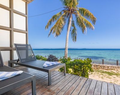 Two lounge chairs with towels sit on a wooden deck overlooking a serene beach with clear blue water and a palm tree under a sunny sky.
