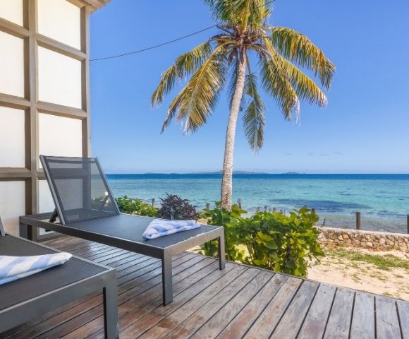 Two lounge chairs with towels sit on a wooden deck overlooking a serene beach with clear blue water and a palm tree under a sunny sky.