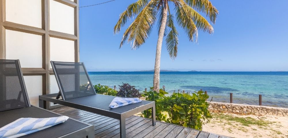 Two lounge chairs with towels sit on a wooden deck overlooking a serene beach with clear blue water and a palm tree under a sunny sky.