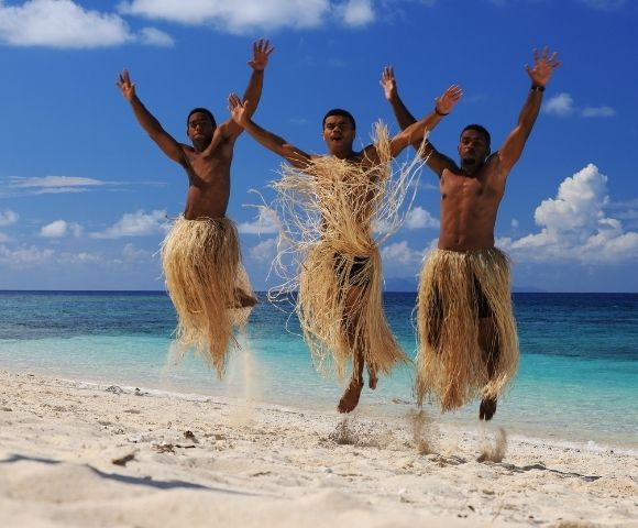 Three people wearing straw skirts joyfully jump on a sunny beach with turquoise sea and clear blue sky in the background, expressing happiness and freedom.
