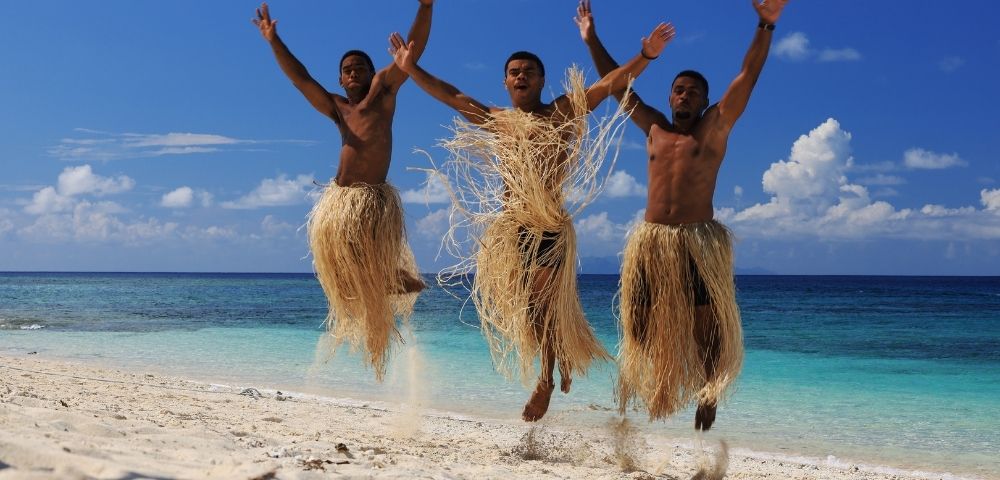 Three people wearing straw skirts joyfully jump on a sunny beach with turquoise sea and clear blue sky in the background, expressing happiness and freedom.