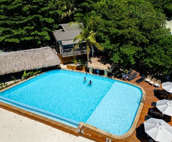 Aerial view of a tropical pool surrounded by lush greenery and thatched huts. Two people swim, while lounge chairs and umbrellas line the wooden deck. Relaxing vibe.