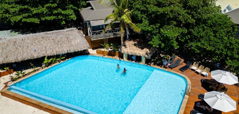 Aerial view of a tropical pool surrounded by lush greenery and thatched huts. Two people swim, while lounge chairs and umbrellas line the wooden deck. Relaxing vibe.
