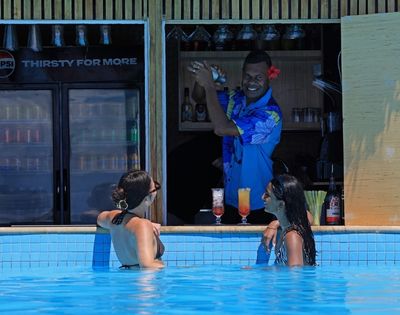 Two women in a pool smile at a bartender mixing drinks at a swim-up bar. The bartender, wearing a blue floral shirt, is cheerful, creating a relaxed, tropical vibe.