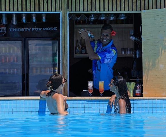 Two women in a pool smile at a bartender mixing drinks at a swim-up bar. The bartender, wearing a blue floral shirt, is cheerful, creating a relaxed, tropical vibe.
