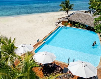 A tropical beach scene with a clear blue pool, sunbathers under white umbrellas, lush palms, a thatched hut, and a calm turquoise sea in the background.