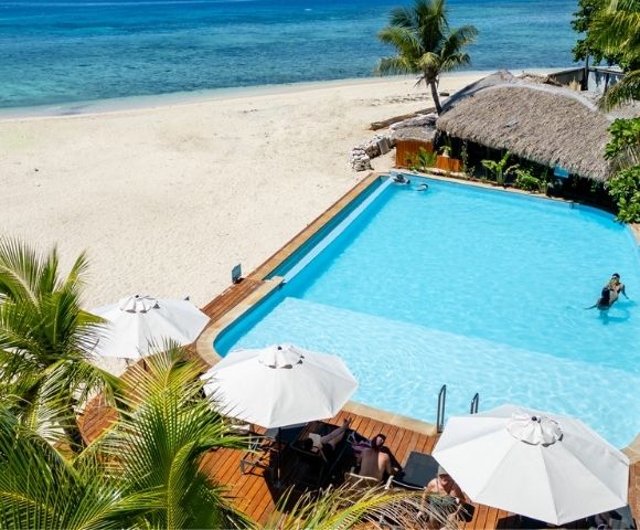 A tropical beach scene with a clear blue pool, sunbathers under white umbrellas, lush palms, a thatched hut, and a calm turquoise sea in the background.