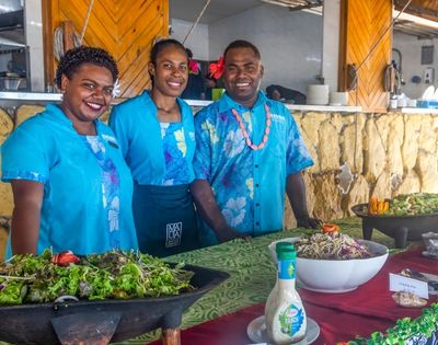 Three people in bright blue shirts smile behind a buffet table with salads and dressings. The setting is cheerful and vibrant, suggesting a festive event.