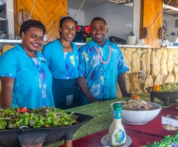 Three people in bright blue shirts smile behind a buffet table with salads and dressings. The setting is cheerful and vibrant, suggesting a festive event.