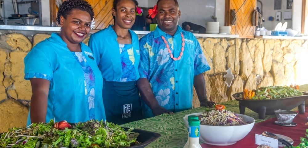 Three people in bright blue shirts smile behind a buffet table with salads and dressings. The setting is cheerful and vibrant, suggesting a festive event.