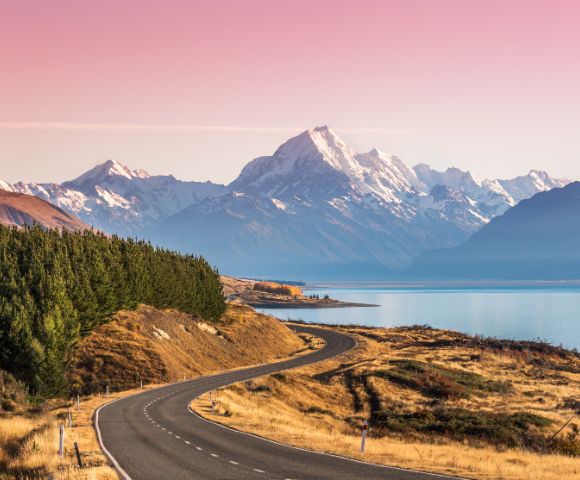 Winding road leads to snow-capped mountains under a pink sky, with a serene lake on the right and dense green trees on the left, evoking tranquility.