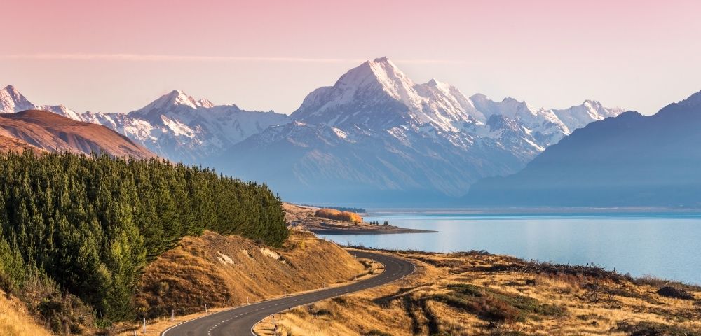 Winding road leads to snow-capped mountains under a pink sky, with a serene lake on the right and dense green trees on the left, evoking tranquility.