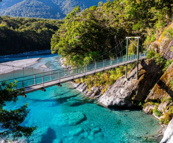 A suspension bridge spans a clear turquoise river, surrounded by lush green foliage and rocky hills, under a bright blue sky, creating a serene atmosphere.