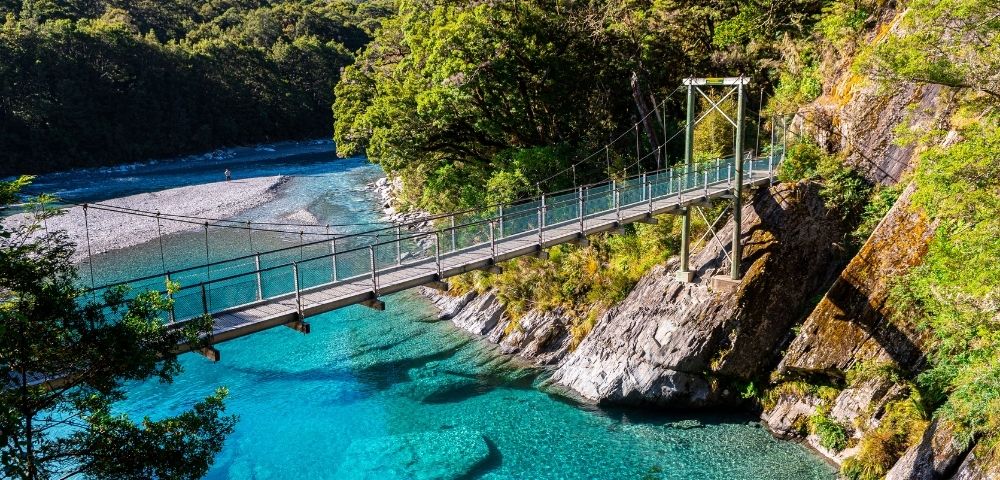 A suspension bridge spans a clear turquoise river, surrounded by lush green foliage and rocky hills, under a bright blue sky, creating a serene atmosphere.
