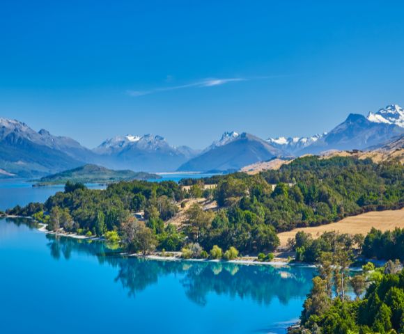 Scenic view of a serene blue lake surrounded by lush green forests and snow-capped mountains under a clear blue sky, conveying tranquility and natural beauty.