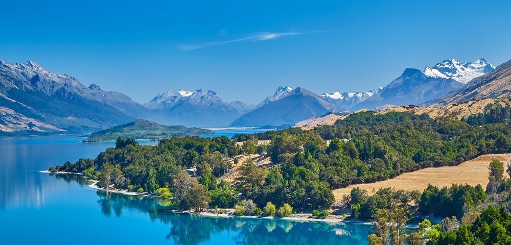 Scenic view of a serene blue lake surrounded by lush green forests and snow-capped mountains under a clear blue sky, conveying tranquility and natural beauty.