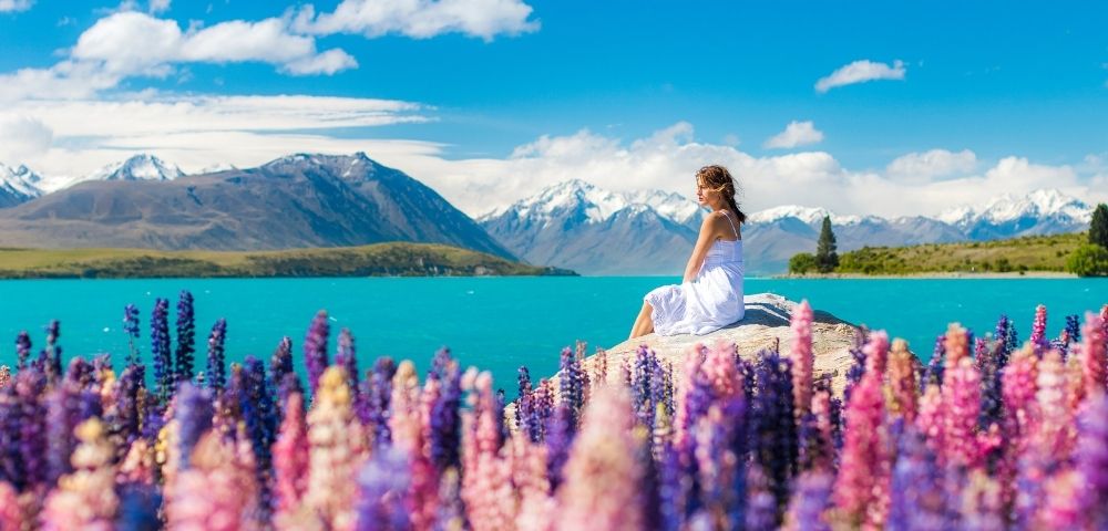 A woman in a white dress sits peacefully on a rock by a turquoise lake, surrounded by vivid purple and pink lupines, with snow-capped mountains in the background.