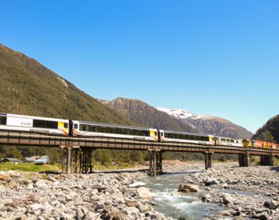 A long passenger train crosses a bridge over a rocky river, framed by green hills and snow-capped mountains under a clear blue sky.