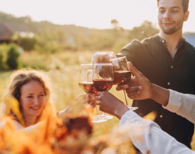 A group of friends clinking wine glasses outdoors at sunset, smiling and celebrating. The setting is warm and cheerful, with a blurry natural background.