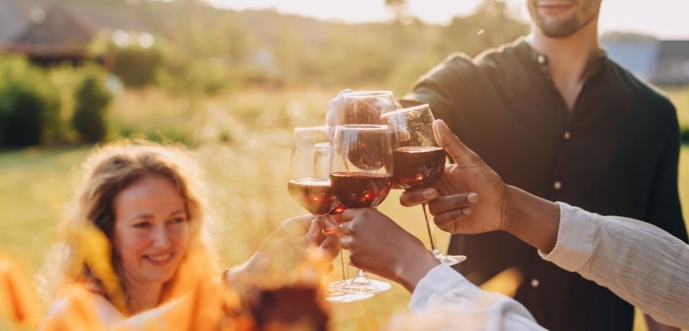 A group of friends clinking wine glasses outdoors at sunset, smiling and celebrating. The setting is warm and cheerful, with a blurry natural background.