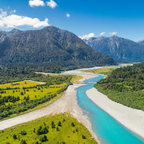 A turquoise river winds through lush green valleys with forested hills and towering mountains in the background under a bright blue sky.