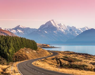 Winding road leads to snow-capped mountains under a pink sky, with a serene lake on the right and dense green trees on the left, evoking tranquility.