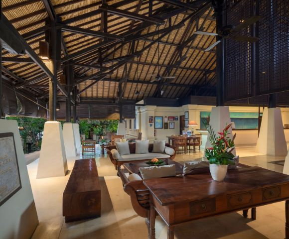 Spacious living area with high, vaulted ceiling and wooden beams. Features elegant furniture, tropical plants, and a warm, inviting ambiance.