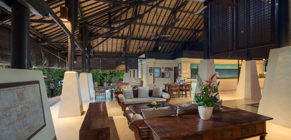 Spacious living area with high, vaulted ceiling and wooden beams. Features elegant furniture, tropical plants, and a warm, inviting ambiance.