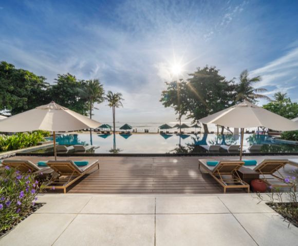 Luxurious poolside scene at sunrise with lounge chairs and umbrellas, overlooking a tranquil infinity pool, lush greenery, and a calm ocean horizon.