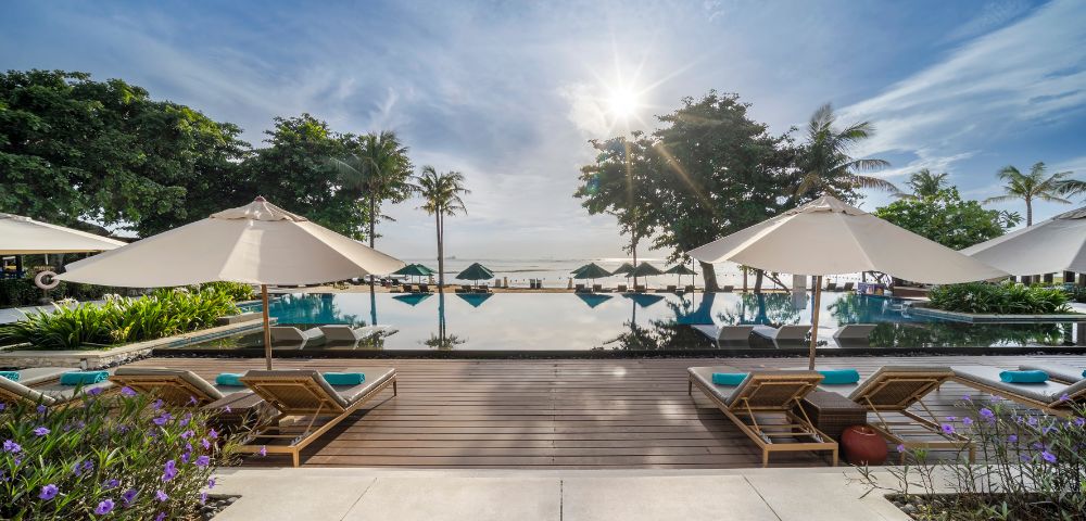 Luxurious poolside scene at sunrise with lounge chairs and umbrellas, overlooking a tranquil infinity pool, lush greenery, and a calm ocean horizon.