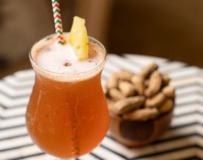 A frothy orange cocktail with a pineapple slice and striped straw sits on a table with a bowl of peanuts in the background, creating a relaxed, tropical vibe.