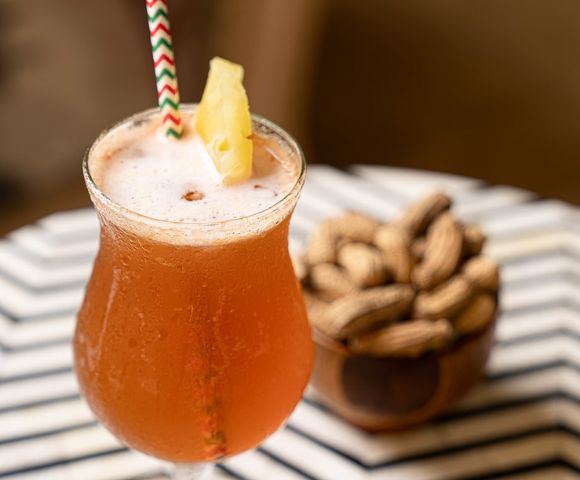 A frothy orange cocktail with a pineapple slice and striped straw sits on a table with a bowl of peanuts in the background, creating a relaxed, tropical vibe.