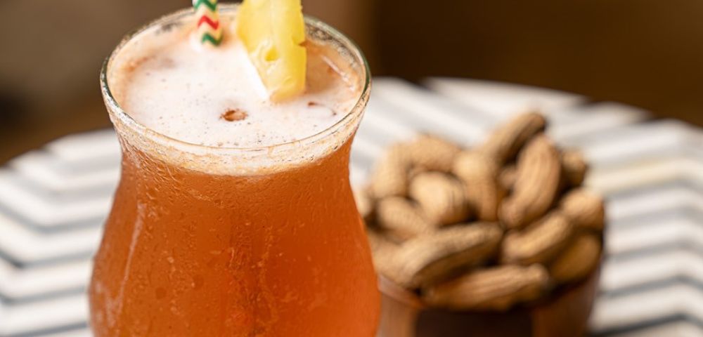 A frothy orange cocktail with a pineapple slice and striped straw sits on a table with a bowl of peanuts in the background, creating a relaxed, tropical vibe.