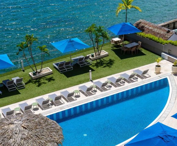 Luxurious seaside pool with blue parasols, white lounge chairs, and palm trees. A serene ocean view enhances the tropical, relaxing ambiance.
