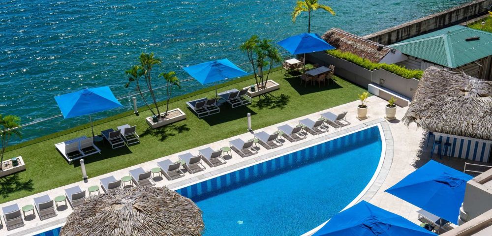 Luxurious seaside pool with blue parasols, white lounge chairs, and palm trees. A serene ocean view enhances the tropical, relaxing ambiance.