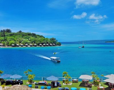 A serene beach scene with vibrant blue water, a boat leaving a wake, distant sailboats, lush greenery, and a clear sky, creating a relaxing tropical atmosphere.